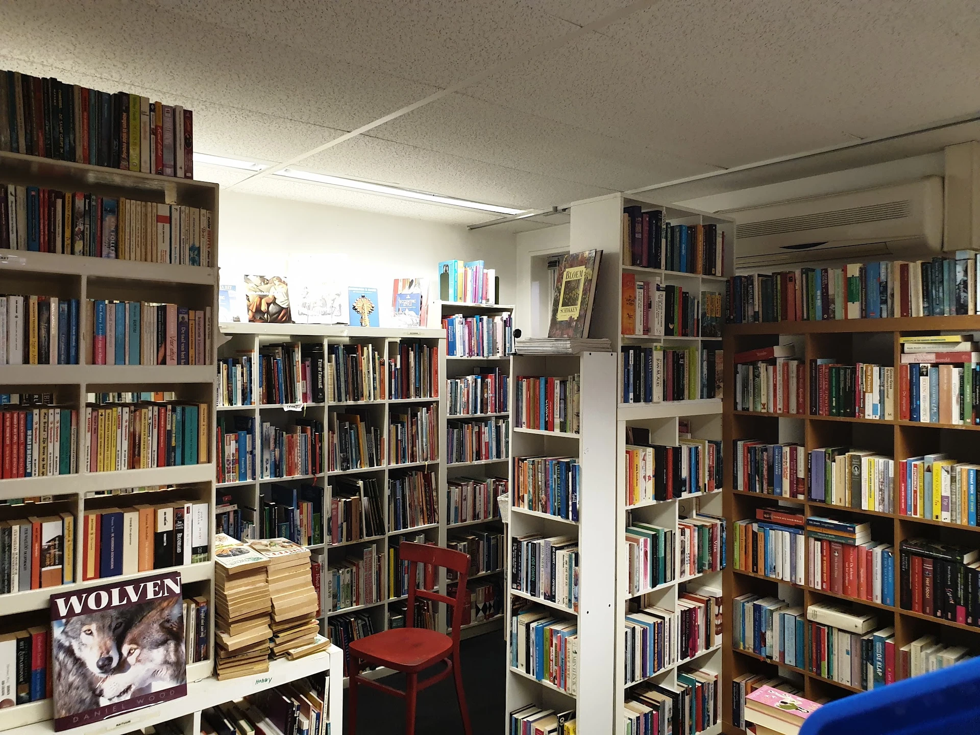 Binnenaanzicht van kringloopwinkel met uitgebreide boekenkasten langs de muren, rode stoel en diverse items op stellingen