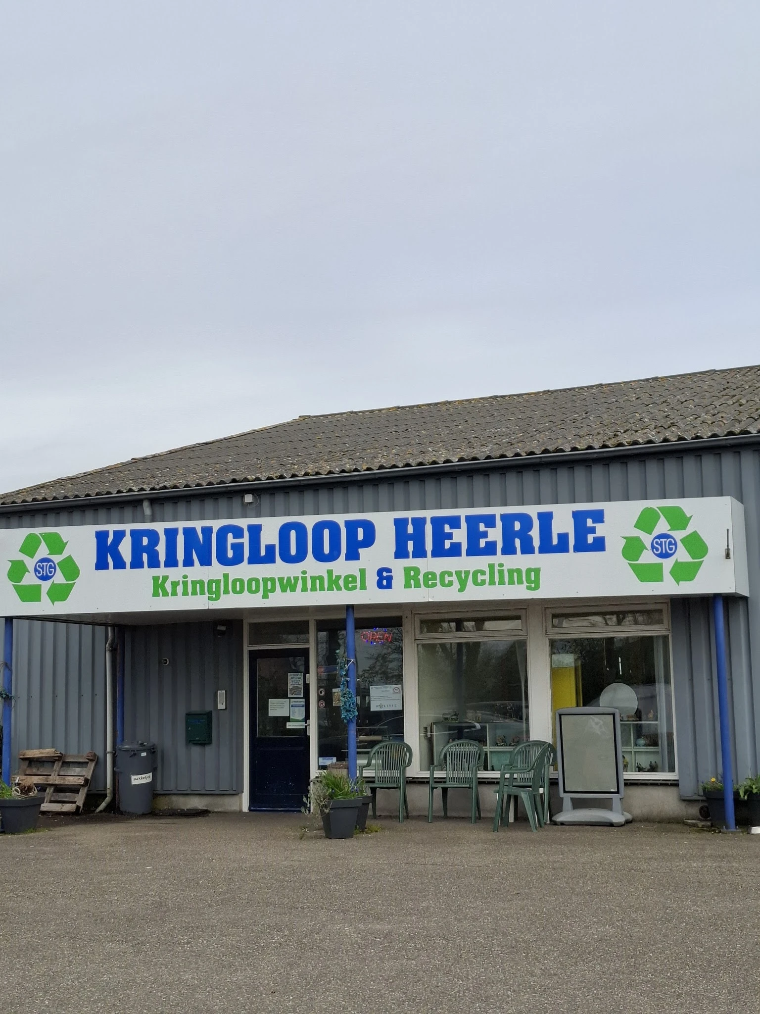Gevel van kringloopwinkel Heerle met blauwe letters en groen recycle-logo's, stellage en ingang zichtbaar