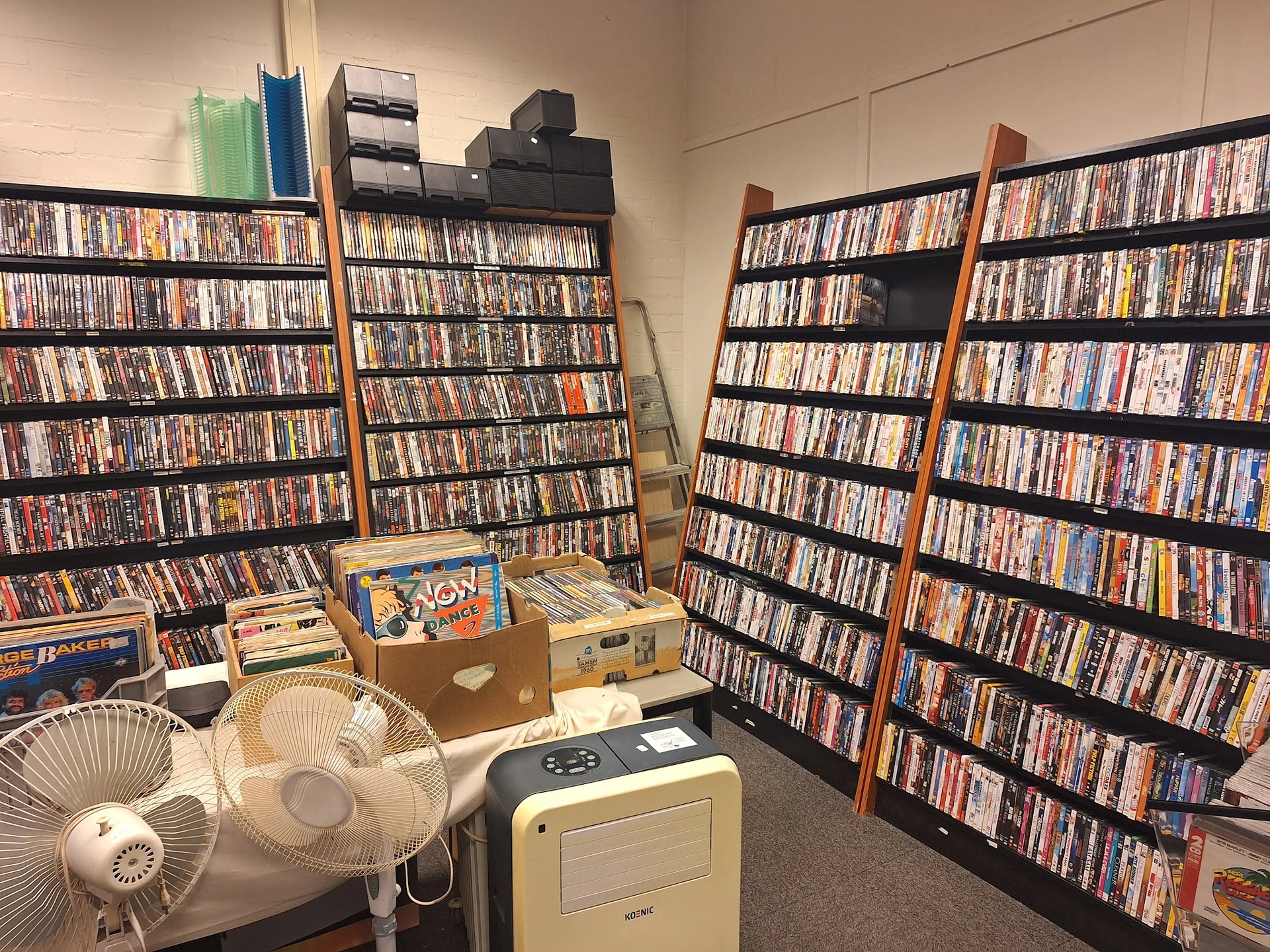 Binnenruimte met meerdere stellingen vol dvd's en cd's, ventilator en kartonnen dozen op de vloer
