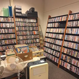 Binnenruimte met meerdere stellingen vol dvd's en cd's, ventilator en kartonnen dozen op de vloer