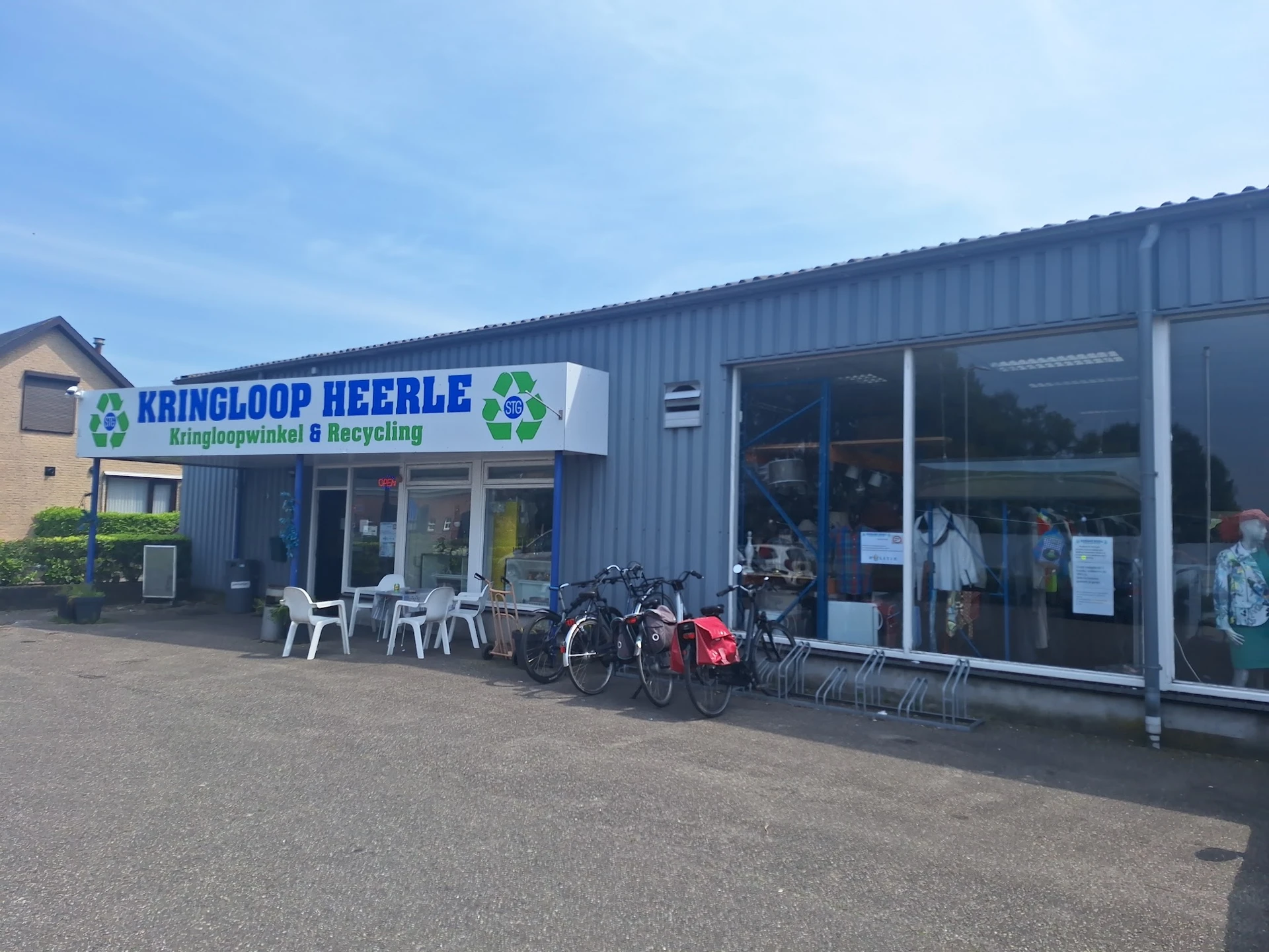 Gevel van Kringloop Heerle met grote witte letters op blauw, ingang met ramen en fietsenstalling ervoor