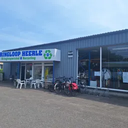 Gevel van Kringloop Heerle met grote witte letters op blauw, ingang met ramen en fietsenstalling ervoor