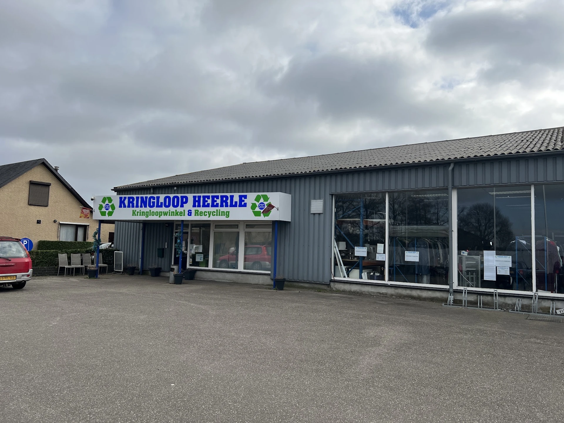 Gevel van kringloopwinkel Heerle met blauwe naam en groene logo's, metalen constructie met grote ramen