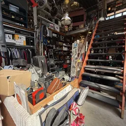 Overzicht van kringloopwinkel met stellingen, dozen en diverse spullen op de vloer en in kasten