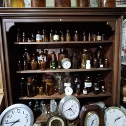 Houten vitrinekast met planken vol antieke glazen flessen, potjes en twee vintage klokken