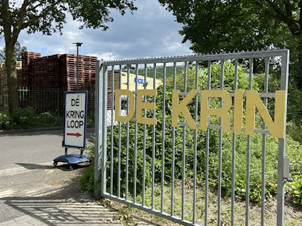 Ingang van kringloopwinkel met grijs hek en geel bord, naambord links zichtbaar