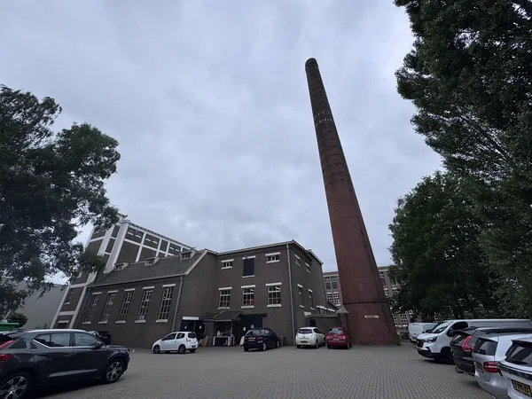 Voorgevel van donker bakstenen gebouw met hoge schoorsteen, parkeerplaats met auto's ervoor