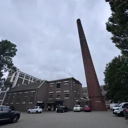 Voorgevel van donker bakstenen gebouw met hoge schoorsteen, parkeerplaats met auto's ervoor