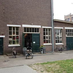 Voorkant van bakstenen kringloopwinkel met groene deuren en ramen, fietsen geparkeerd ervoor
