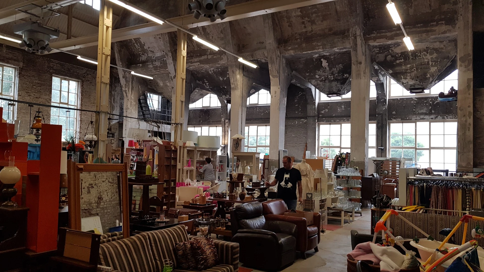 Ruime kringloopwinkel met diverse meubels, boeken en spullen op stellingen en tafels, grote ramen en industrieel plafond