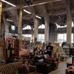 Ruime kringloopwinkel met diverse meubels, boeken en spullen op stellingen en tafels, grote ramen en industrieel plafond