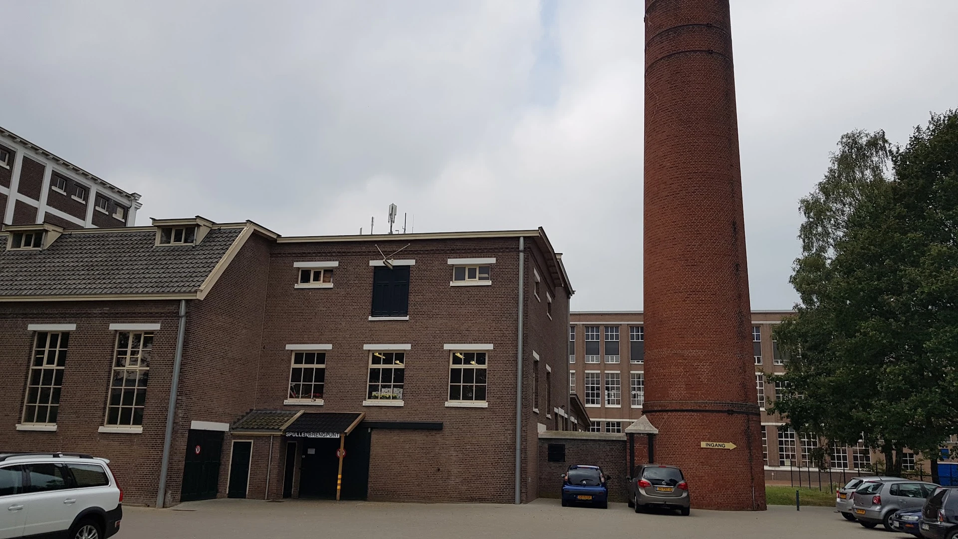 Voorgevel van een bakstenen kringloopwinkel met grote ramen, twee auto's ervoor geparkeerd en een industriële schoorsteen ern