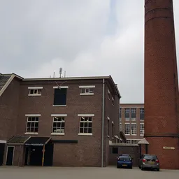Voorgevel van een bakstenen kringloopwinkel met grote ramen, twee auto's ervoor geparkeerd en een industriële schoorsteen ern