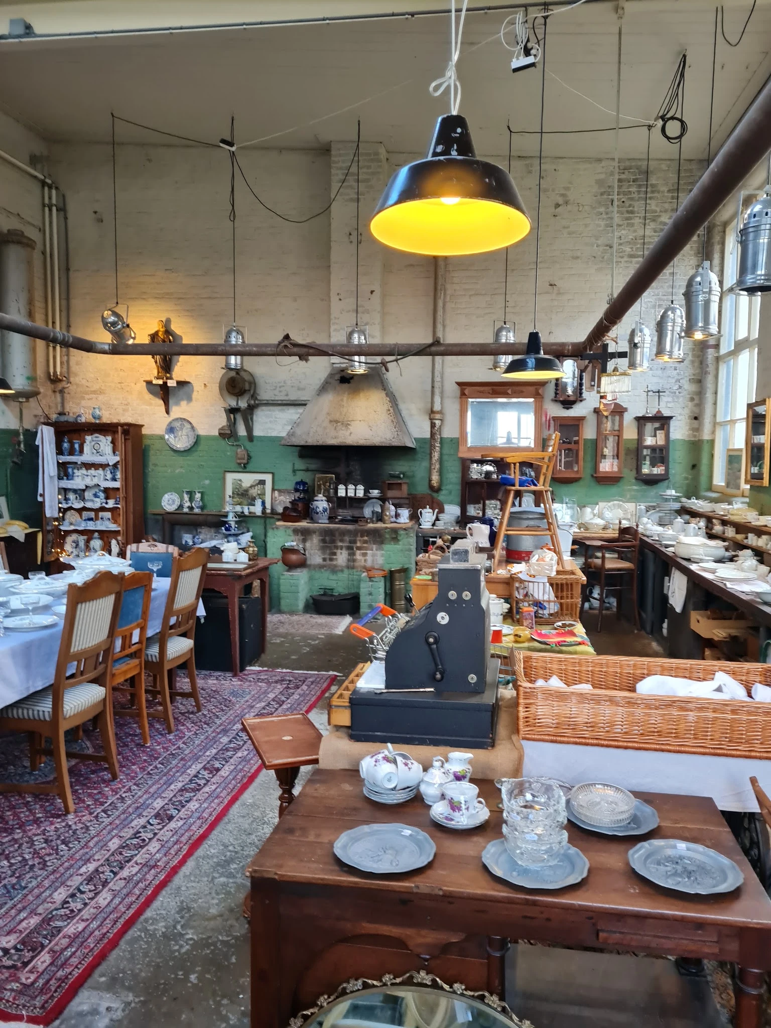 Ruim interieur van kringloopwinkel met groene muren, hanglampen, diverse meubels en spullen op tafels en rekken