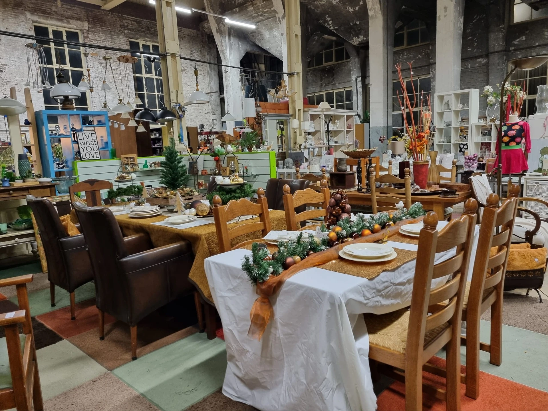Industriële kringloopwinkel met opgezet banketdiner: houten stoelen, witte tafelkleden, groenendecoratie en serviesgoed op la