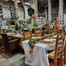 Industriële kringloopwinkel met opgezet banketdiner: houten stoelen, witte tafelkleden, groenendecoratie en serviesgoed op la