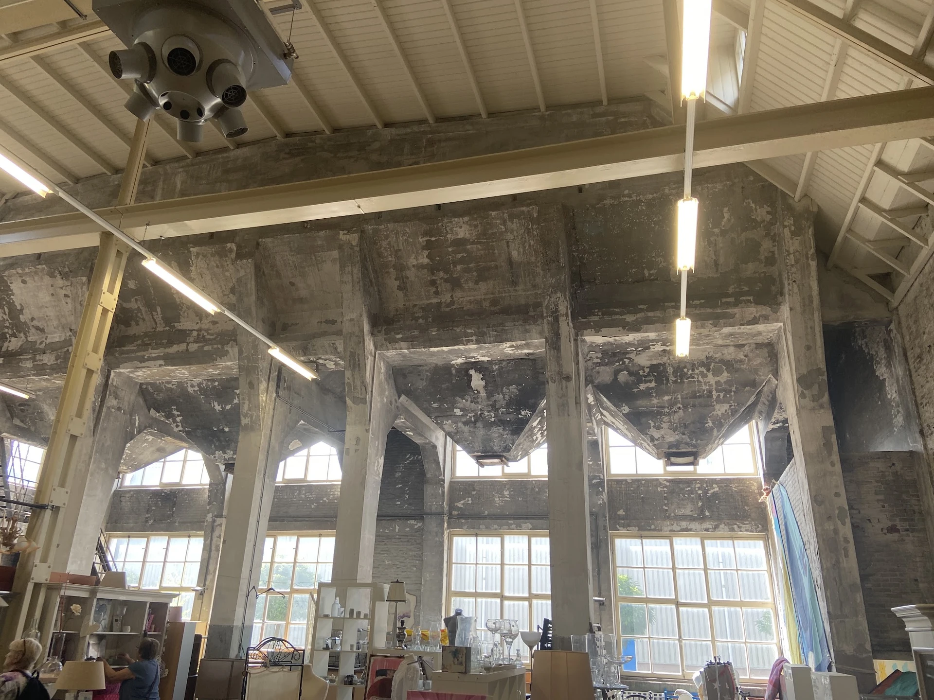 Ruim interieur van kringloopwinkel met hoge plafonds, grote ramen, verlichting en enkele bezoekers