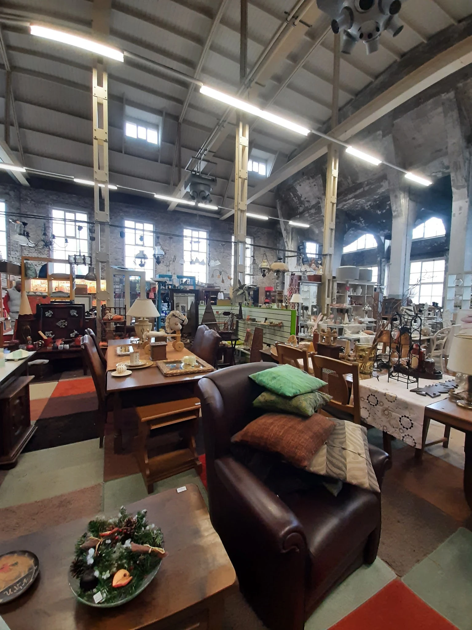 Ruim interieur van kringloopwinkel met tweedehands meubels, stoelen, tafels en kussens uitgestald tussen grote raampartijen