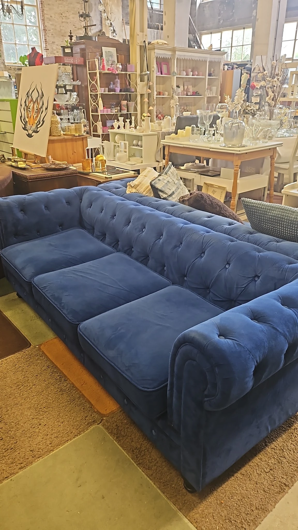 Blauwe chesterfield-bank met kapitonnering en houten poten, tentoongesteld in een kringloopwinkel