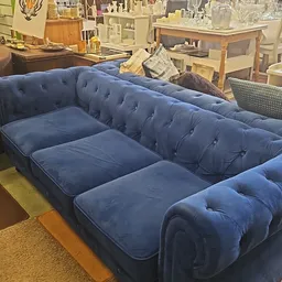 Blauwe chesterfield-bank met kapitonnering en houten poten, tentoongesteld in een kringloopwinkel
