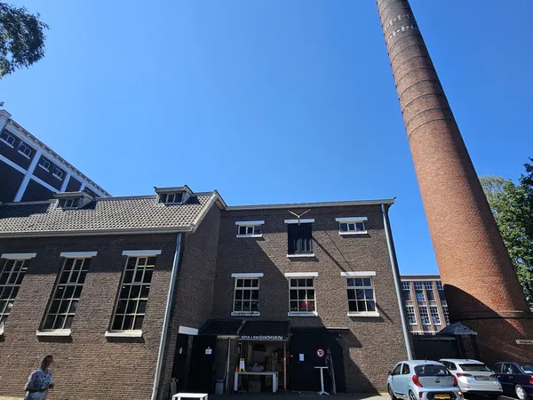 Bakstenen voorgevel van kringloopwinkel Het Ketelhuis met zwarte ingang en ramen, naast hoge rode schoorsteen