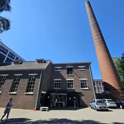Bakstenen voorgevel van kringloopwinkel Het Ketelhuis met zwarte ingang en ramen, naast hoge rode schoorsteen
