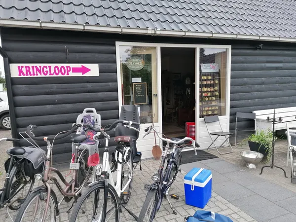 Voorkant kringloopwinkel met grijze garagedeur, witte deur, roze pijl met 'KRINGLOOP', fietsen en blauwe koelbox op terras