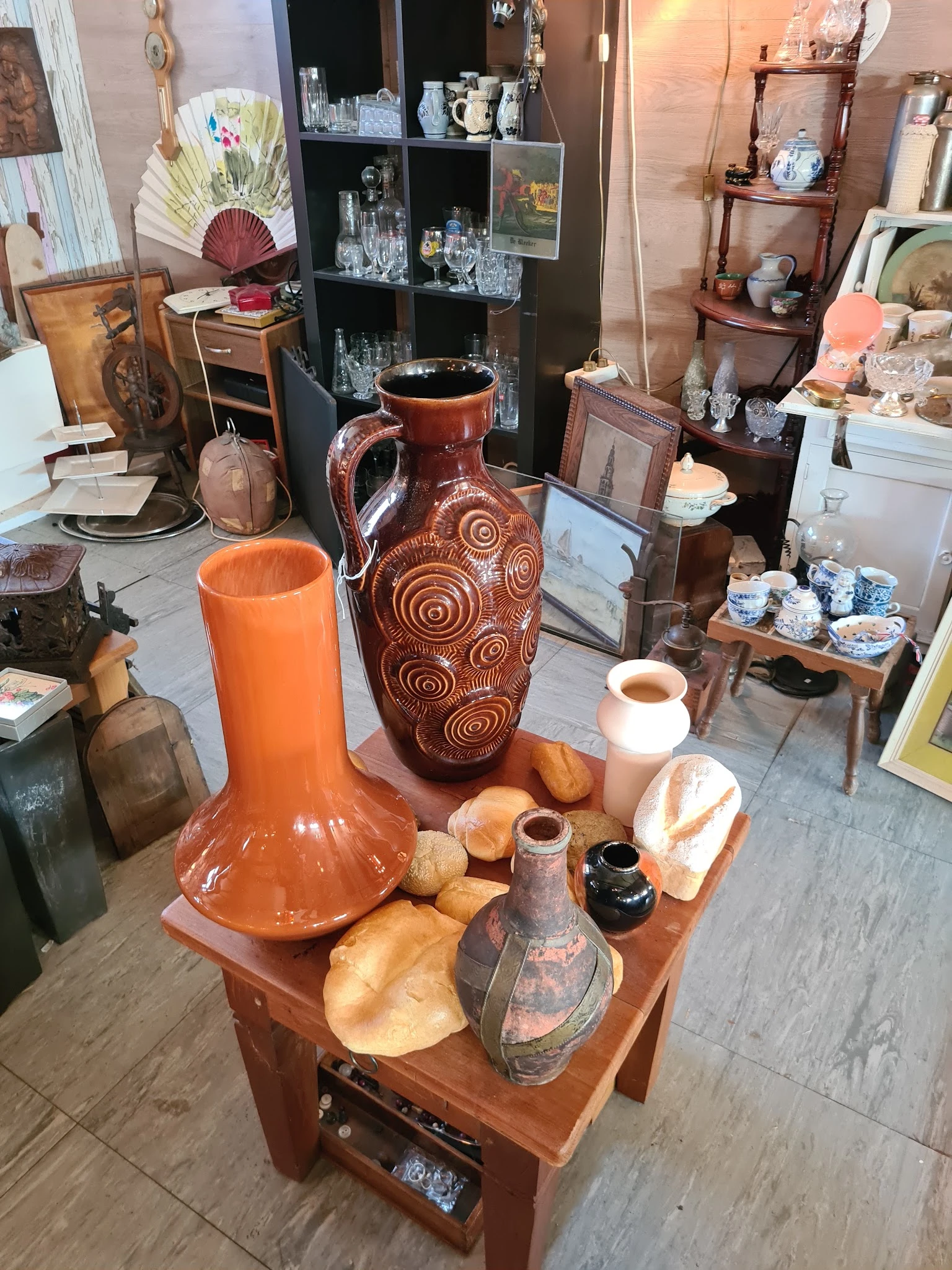 Uitgestalde keramische en aardewerk items op een houten tafel, waaronder oranje vazen, decoratieve potten en andere huishoude