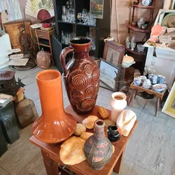 Uitgestalde keramische en aardewerk items op een houten tafel, waaronder oranje vazen, decoratieve potten en andere huishoude