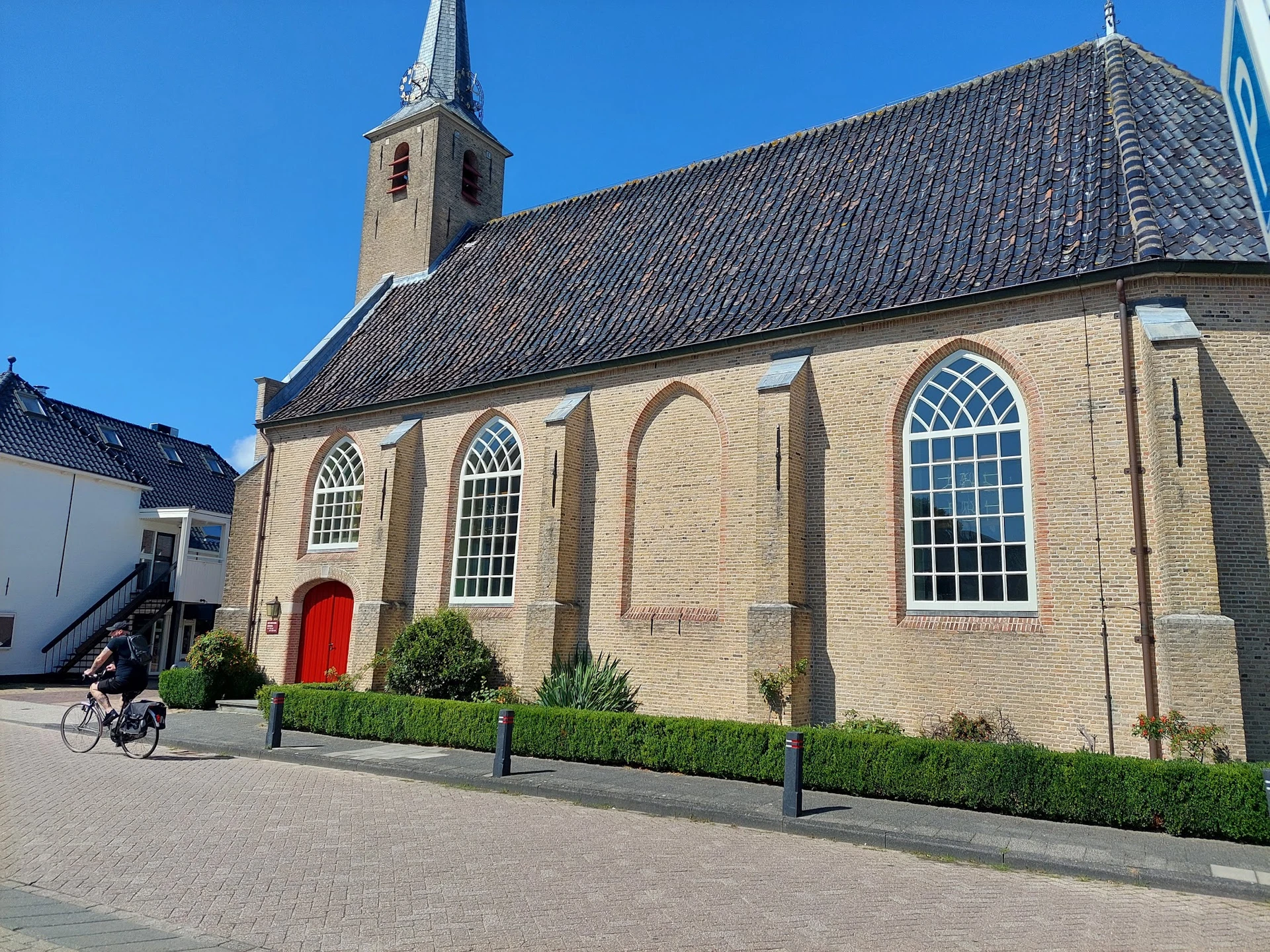 Voorgevel van een bakstenen kerk met rode deur, grote ramen en toren tegen blauwe lucht