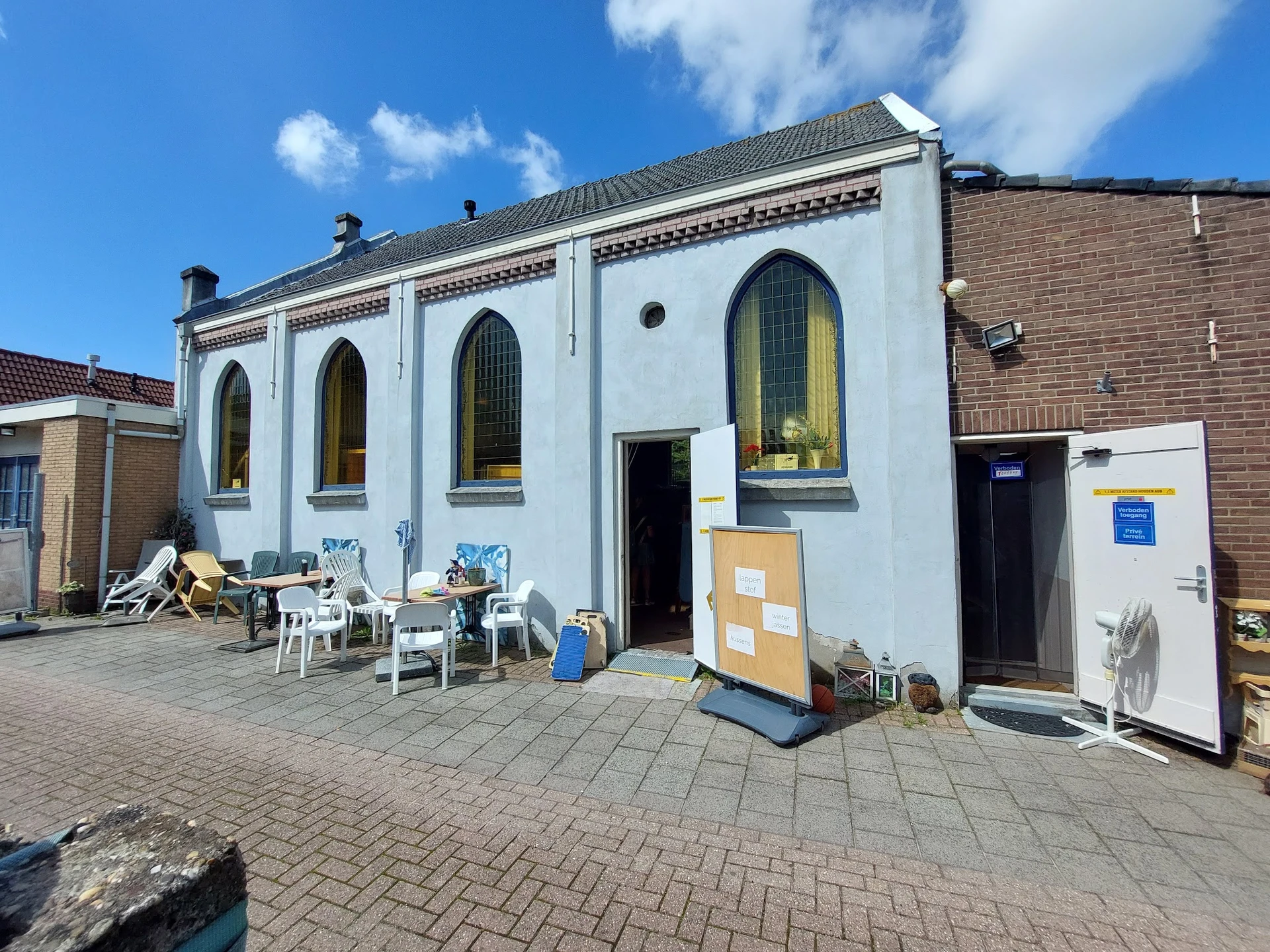 Witte gevel van kringloopwinkel met grote ramen, terras met stoelen ervoor en bakstenen straatwerk