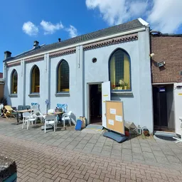 Witte gevel van kringloopwinkel met grote ramen, terras met stoelen ervoor en bakstenen straatwerk
