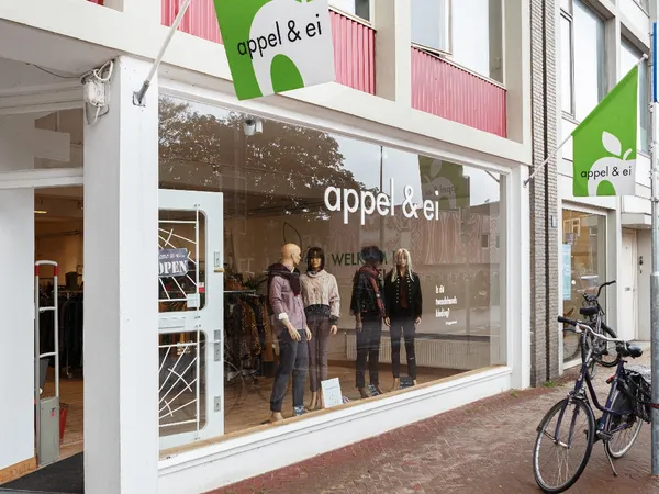Winkelgevel van kringloopwinkel met grote glazen pui, groene en roze belettering, etalagepop en klanten zichtbaar