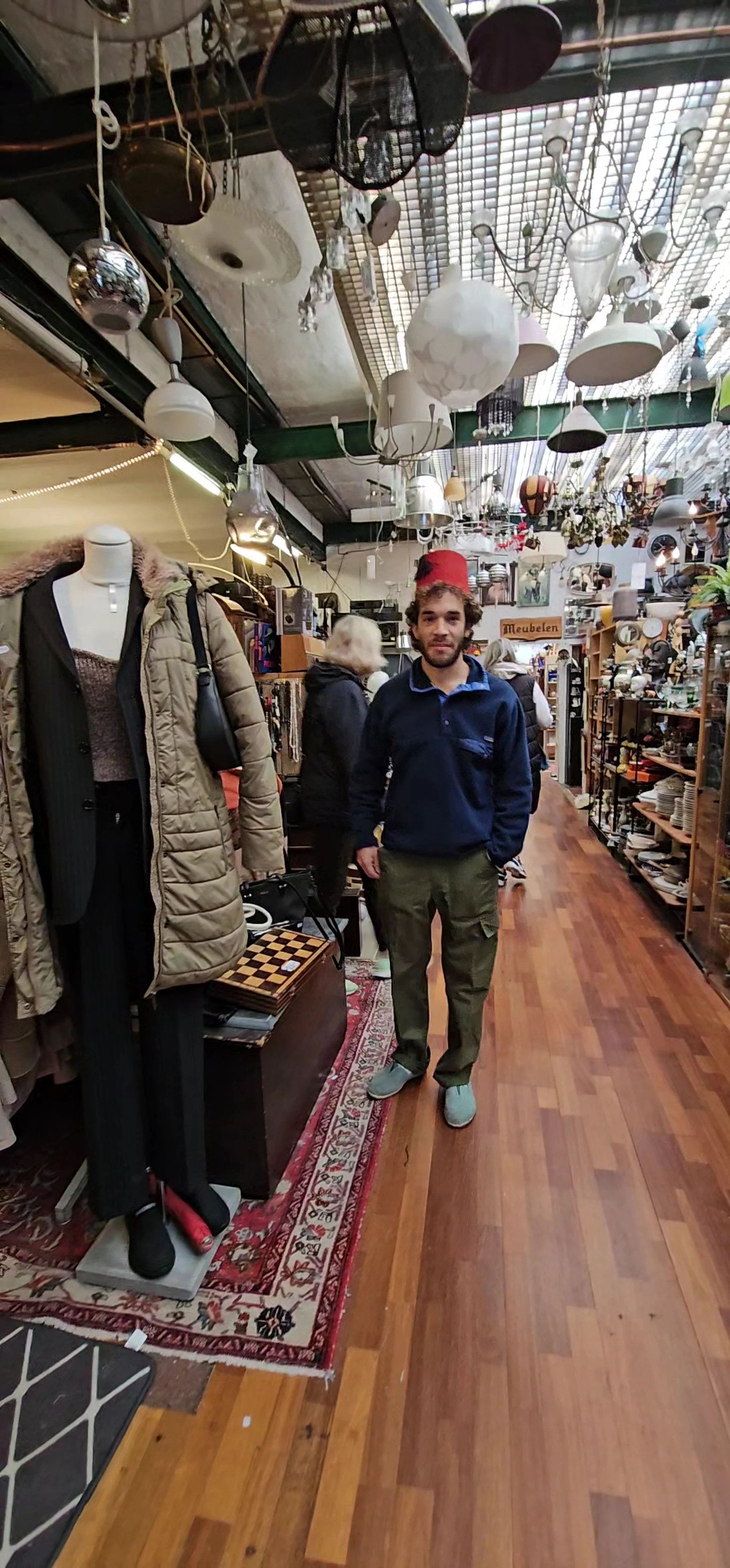 Man in rode pet en blauwe trui in kringloopwinkel met kleding en spullen op achtergrond