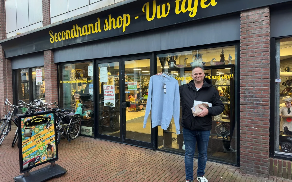 Voorgevel van tweedehandswinkel met grote ramen, gele letters op zwarte band, man in zwarte jas voor ingang