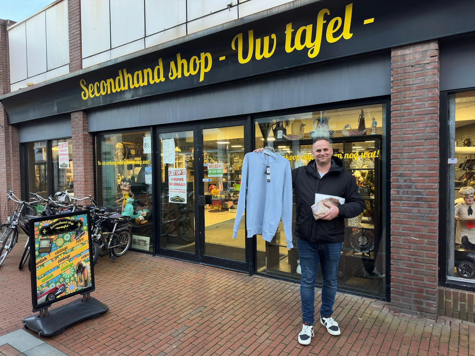 Voorgevel van tweedehandswinkel met grote ramen, gele letters op zwarte band, man in zwarte jas voor ingang