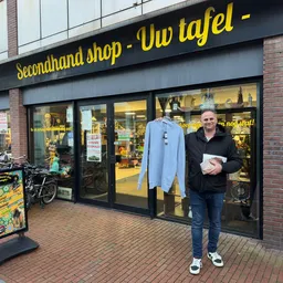 Gevel van secondhandwinkel met grote ramen, gele letters op donker bord, man voor ingang met artikel