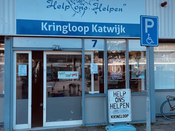 Blauwe gevel van kringloopwinkel met nummer 7, ramen met uitgestalde spullen en 'Help ons Helpen' borden