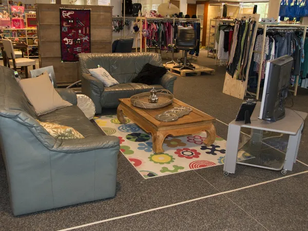 Zithoek met grijze banken en houten tafel in kringloopwinkel, achtergrond met kledingrekken