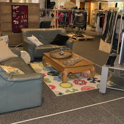 Zithoek met grijze banken en houten tafel in kringloopwinkel, achtergrond met kledingrekken