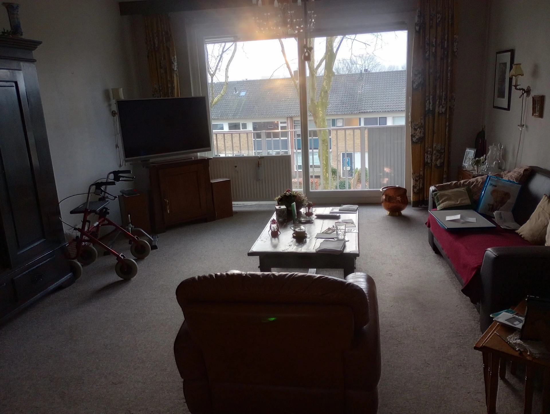Woonkamer met bruine fauteuil, TV op meubel, witte deur naar balkon en diverse meubels