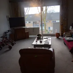 Woonkamer met bruine fauteuil, TV op meubel, witte deur naar balkon en diverse meubels