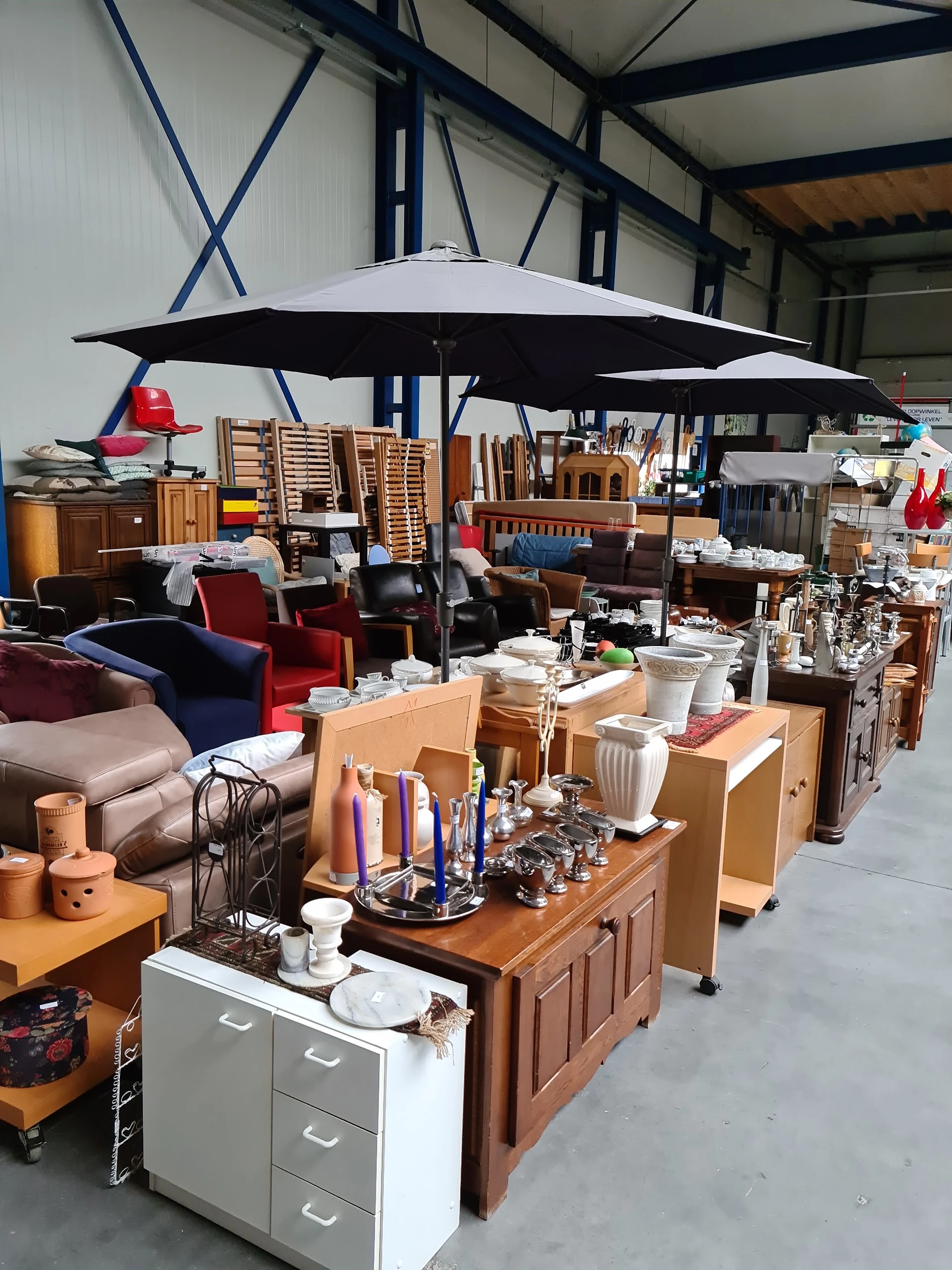 Overzicht van kringloopwinkelinterieur met meubels, serviesgoed, huishoudspullen en zwarte parasols boven winkelgedeelte