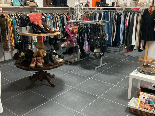 Binnenaanzicht kringloopwinkel met kledingrekken, vintage tafel met schoenen en diverse tweedehands spullen op donkere tegelv