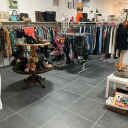 Binnenaanzicht kringloopwinkel met kledingrekken, vintage tafel met schoenen en diverse tweedehands spullen op donkere tegelv