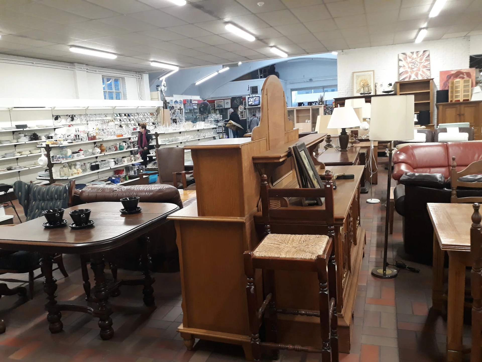 Winkelvloer met tweedehands meubels: tafels, stoelen, kasten en een roze bank; op de achtergrond stellingen met spullen