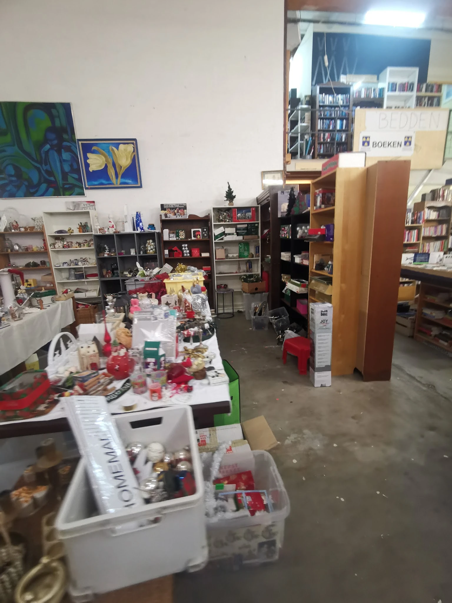 Binnenzicht van kringloopwinkel met tafels vol spullen, stellingen en dozen verspreid over de winkelvloer
