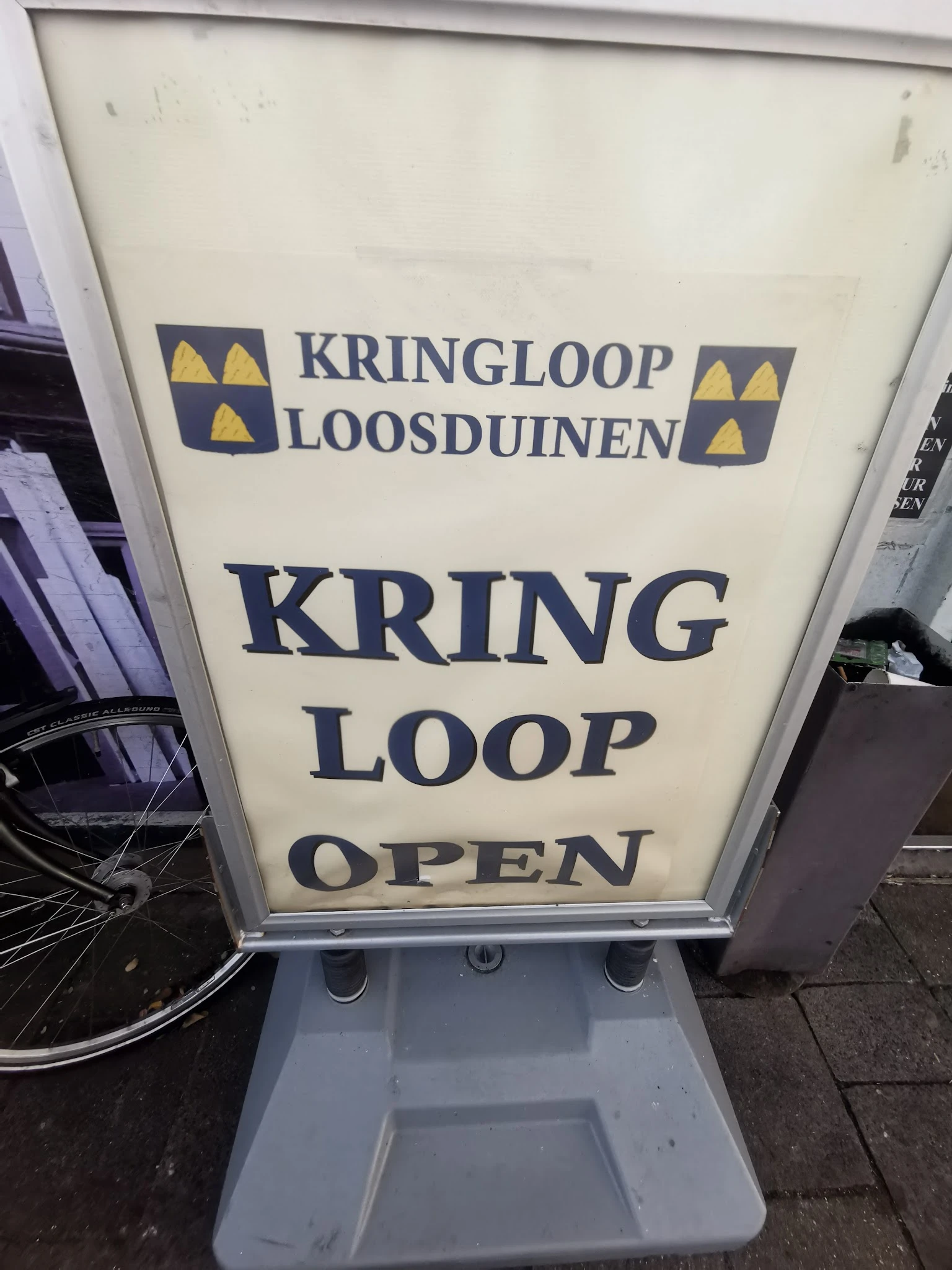 Wit A-bord met donkerblauwe tekst 'KRING LOOP OPEN' en logo van Kringloop Loosduinen op straat