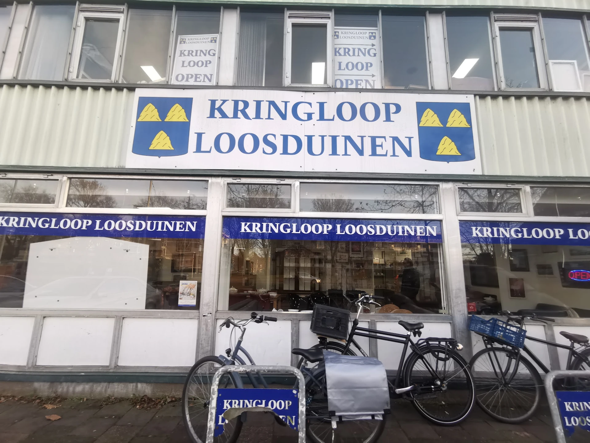 Voorkant van kringloopwinkel met blauwe gevel en gele driehoek-logo's, fietsen geparkeerd voor de etalage