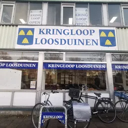 Voorkant van kringloopwinkel met blauwe gevel en gele driehoek-logo's, fietsen geparkeerd voor de etalage